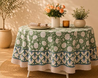 Può includere: Una tovaglia rotonda caratterizzata da un motivo floreale verde e bianco sulla parte superiore, con un bordo smerlato blu e verde e un orlo a righe marroni e bianche. Il tessuto copre un tavolo rotondo, creando un aspetto stratificato e definito.