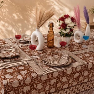 May include: A brown and white floral patterned tablecloth with matching placemats and napkins. The table is set with white circular vases, a brown glass bottle, red and blue drinks in clear glasses, and a bouquet of red and white flowers.