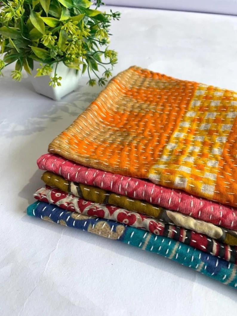 May include: A stack of five folded fabric textiles featuring various patterns and colors including orange, yellow, pink, olive green, red, and blue. Each piece of fabric is stitched with visible white thread running in parallel lines.
