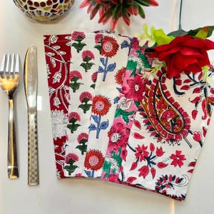K&ouml;nnte beinhalten: Eine Sammlung farbenfroher Servietten mit Blumendruck in Rot, Rosa und Wei&szlig;. Die Servietten sind mit einer Gabel und einem Messer angeordnet, und ein dekorativer Kerzenhalter und Blumen befinden sich im Hintergrund. Die Servietten weisen verschiedene Blumenmuster auf.