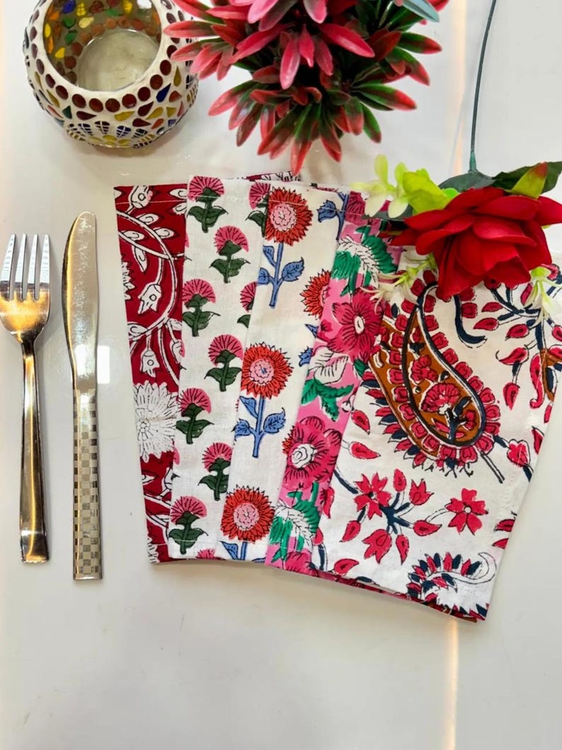 K&ouml;nnte beinhalten: Eine Sammlung farbenfroher Servietten mit Blumendruck in Rot, Rosa und Wei&szlig;. Die Servietten sind mit einer Gabel und einem Messer angeordnet, und ein dekorativer Kerzenhalter und Blumen befinden sich im Hintergrund. Die Servietten weisen verschiedene Blumenmuster auf.