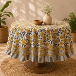 Pu&ograve; includere: Una tovaglia rotonda con base bianca e un motivo floreale giallo ripetuto, un'ampia bordura decorativa con intricati motivi botanici grigi e gialli e un sottile bordo a righe. Una piccola ciotola di legno e un vaso con fiori sono appoggiati sopra.