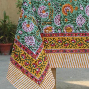 May include: A green floral tablecloth with a repeating pattern of pink and orange flowers. The border features a yellow and red floral design with white and yellow striped edges. The tablecloth is draped over a wooden table.