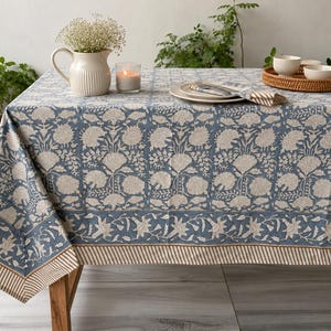 Puede incluir: Un mantel rectangular que presenta un patrón de estampado floral en azul y beige con una disposición de cuadrícula. La tela tiene un borde ancho con un motivo floral más pequeño y un borde fino a rayas. El diseño cubre toda la superficie de la mesa.
