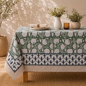 May include: A rectangular tablecloth featuring a green and white floral pattern on the top surface, with a decorative border of blue and white fan-shaped motifs and a thin striped edge in beige and white, draped over a wooden table.