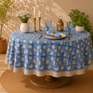 Pu&ograve; includere: Una tovaglia rotonda con un motivo floreale blu e bianco e un bordo a righe. Il tessuto copre un tavolo circolare, apparecchiato con un vaso bianco di margherite, due candelabri in ottone, bicchieri blu e un tovagliolo.