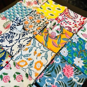 K&ouml;nnte beinhalten: Eine Sammlung bunter Stoffservietten mit floralen Mustern. Die Servietten zeigen verschiedene Designs, darunter rosa, orange und blaue Blumen, die in einem Raster angeordnet sind. Einige Servietten sind gefaltet, um ihre Muster zu zeigen.