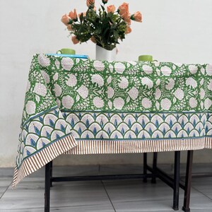Puede incluir: Un mantel verde con estampado floral y borde decorativo, sobre una mesa de metal negro. Un jarr&oacute;n con flores color melocot&oacute;n y dos tazas verdes est&aacute;n sobre la mesa. El mantel tiene un patr&oacute;n floral repetido.