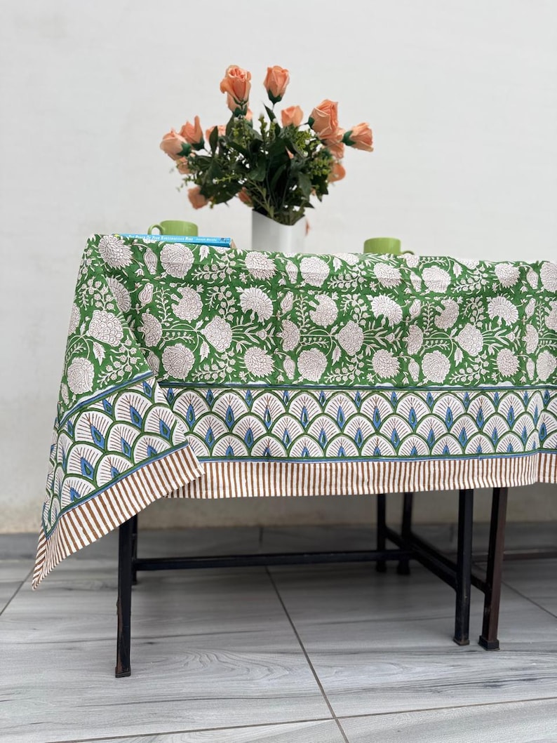 Puede incluir: Un mantel verde con estampado floral y borde decorativo, sobre una mesa de metal negro. Un jarr&oacute;n con flores color melocot&oacute;n y dos tazas verdes est&aacute;n sobre la mesa. El mantel tiene un patr&oacute;n floral repetido.