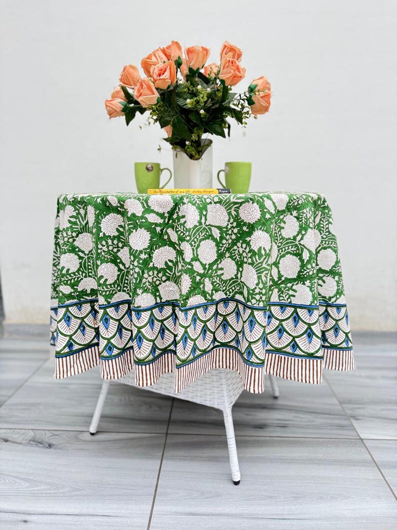 Pu&ograve; includere: Un tavolo rotondo coperto da una tovaglia con stampa floreale verde e bianca, caratterizzata da un bordo smerlato blu e marrone. Sul tavolo si trova un vaso bianco riempito di rose color pesca e due tazze verde chiaro.