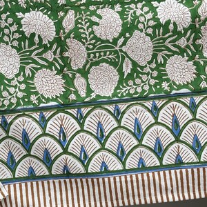 Puede incluir: Textil con estampado floral verde y blanco. El dise&ntilde;o presenta grandes flores blancas y follaje sobre un fondo verde. El borde tiene un patr&oacute;n en forma de abanico con detalles azules y rayas verticales marrones.