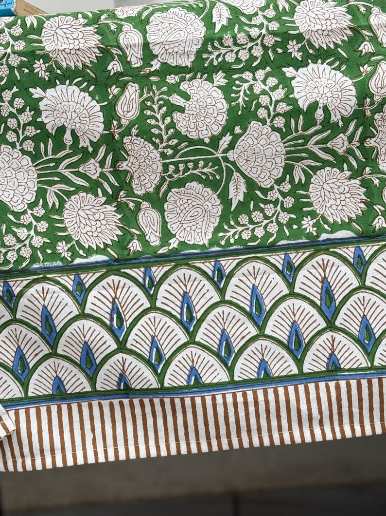 Puede incluir: Textil con estampado floral verde y blanco. El dise&ntilde;o presenta grandes flores blancas y follaje sobre un fondo verde. El borde tiene un patr&oacute;n en forma de abanico con detalles azules y rayas verticales marrones.