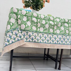 Puede incluir: Un mantel verde con un estampado floral blanco y un borde decorativo. El borde presenta un dise&ntilde;o repetitivo de formas azules y verdes con rayas marrones. Un jarr&oacute;n de flores color melocot&oacute;n est&aacute; sobre la mesa.
