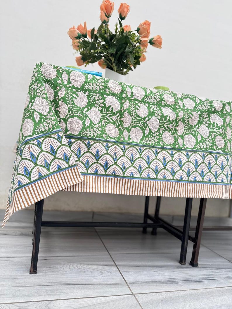 Puede incluir: Un mantel verde con un estampado floral blanco y un borde decorativo. El borde presenta un dise&ntilde;o repetitivo de formas azules y verdes con rayas marrones. Un jarr&oacute;n de flores color melocot&oacute;n est&aacute; sobre la mesa.
