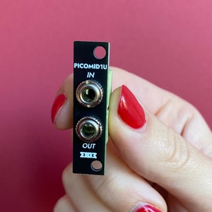 Op de afbeelding: Een zwarte PICOMID1U MIDI-interfacemodule met twee 6,35 mm jacks met de labels "IN" en "OUT".
