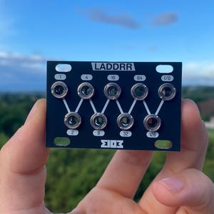 Op de afbeelding: Zwart elektronisch module met de tekst "LADDRR" en genummerde input/output jacks. De module wordt vastgehouden tegen een achtergrond van een blauwe lucht en groen landschap.