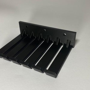 Pode incluir: Prateleira de parede de plástico preto com cinco ganchos. A prateleira é projetada para segurar itens como chaves, toalhas ou outros objetos pequenos.