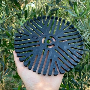 Pode incluir: Um objeto preto, circular, com um orifício central e raios radiais. O objeto é segurado em uma mão contra um fundo de folhagem verde. O objeto tem o texto "SITE".