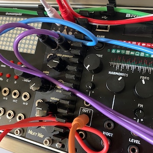 May include: Close-up of a black synthesizer module with various colored cables plugged in. The module has knobs, ports, and text including "Milky Way," "Harmonics," and "Out 1." Cables are red, white, blue, purple, and orange.