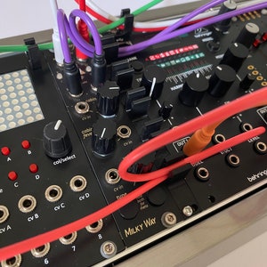 May include: A close-up of a black and silver synthesizer module with red, white, purple and green cables plugged in. The module has knobs, buttons and jacks. The text "MILKY WAY" is printed on the module.