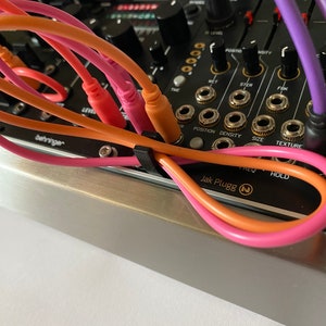 May include: A close-up of a black synthesizer with various knobs and buttons.  The synthesizer has orange and pink patch cables connected to it. The text "Jak Plugg" is visible on the synthesizer.
