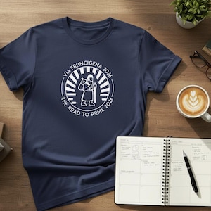 Op de afbeelding: Marineblauw t-shirt met een wit cirkelvormig ontwerp. Het ontwerp zegt "VIA FRANCIGENA 2026 THE ROAD TO ROME 2026" en toont een pelgrim. Het shirt ligt op een houten oppervlak met een notitieboek en koffie.