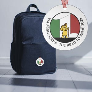 Op de afbeelding: Een donkerblauwe rugzak met een ronde versiering met de kleuren van de Italiaanse vlag en een pelgrimillustratie. Het ornament luidt "VIA FRANCIGENA THE ROAD TO ROME 2026."