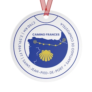 Op de afbeelding: Een rond wit keramisch ornament met een rood lint. Het ontwerp toont een blauwe kaart van de Camino Frances-route, een gouden sint-jakobsschelp en tekst waaronder 790 km, 2.591.864 ft en Saint-Jean-Pied-de-Port naar Santiago de Compostela.