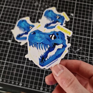 Pode incluir: Um adesivo azul e branco com um crânio de dinossauro com o texto "先史時代" e o nome de usuário do artista nas redes sociais "CHARRYDRAWGOOD".