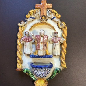 Puede incluir: Una placa de pared de cerámica con un tema religioso. Presenta una figura central con una túnica blanca y una faja roja, rodeada de ángeles y una cruz. La placa tiene un cuenco azul y blanco en la parte inferior.