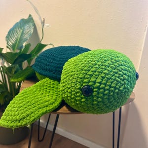 Pode incluir: Uma tartaruga de pelúcia feita à mão, em tons de verde. A tartaruga apresenta um corpo texturizado de crochê, com uma cabeça e barbatanas verde brilhante e uma carapaça verde escura. Olhos pretos de botão adicionam detalhes. A tartaruga está numa mesa redonda.