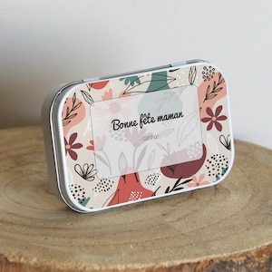 May include: A white metal tin with a floral design in shades of pink, red, and green. The tin has a clear label with the text "Bonne fete maman -GINNY-"