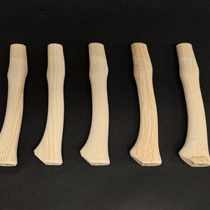 Könnte beinhalten: Fünf unfertige Axtgriffe aus Holz, alle gleich groß und gleich geformt, sind in einer Reihe auf einer schwarzen Oberfläche angeordnet.