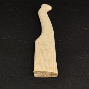 Könnte beinhalten: Ein unlackierter, leicht gebogener Axt-Stiel aus Holz. Der Stiel ist aus hellem Holz und hat eine glatte Oberfläche.