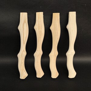 Könnte beinhalten: Vier unfertige Holzmöbelfüße mit einem natürlichen Holzmaserungsmuster. Jeder Fuß hat ein geschwungenes Design, das oben und unten breiter und in der Mitte schmaler ist. Die Beine sind hellbraun und vor einem schwarzen Hintergrund platziert.