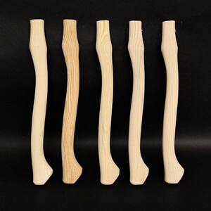 Könnte beinhalten: Fünf unfertige Axtgriffe aus Holz, alle mit einer ähnlichen gebogenen Form. Die Griffe sind hellbraun und haben eine glatte, polierte Oberfläche.