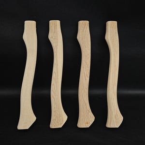 Könnte beinhalten: Vier unfertige Axt-Stiele aus Holz, jeder mit einer leicht unterschiedlichen Krümmung und Maserung. Die Stiele sind hellbraun und scheinen aus Hartholz zu sein.