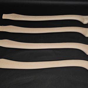 Könnte beinhalten: Vier unfertige Axt-Stiele aus Holz, alle gleich groß und in der gleichen Form, sind in einer Reihe auf einer schwarzen Oberfläche angeordnet.