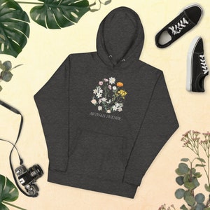 Puede incluir: Una sudadera con capucha gris oscuro con un diseño floral y el texto "Artisan Avenue" impreso en la parte delantera.