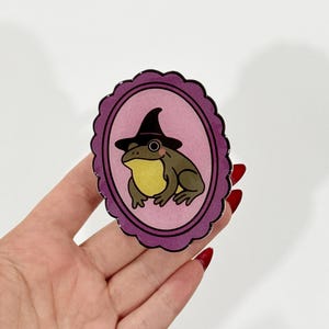 May include: A sticker featuring a cartoon frog wearing a witch hat, set against a pink oval background with a scalloped purple border. The frog is green and yellow, with a black hat. The sticker is held by a hand.