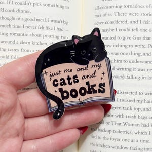 May include: A black cat-shaped sticker with the text "just me and my cats and books" on a light pink background. The sticker is held in a hand over an open book.