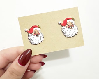 May include: A pair of Santa Claus face earrings. Each earring features a red hat, white beard, and rosy cheeks. The earrings are on a tan card, held by a hand with red nail polish. The earrings are a festive accessory.