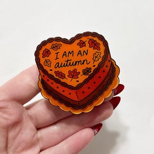 May include: A heart-shaped sticker with the text "I AM AN autumn" in orange and brown. The sticker has a cake-like design with autumn leaves and a scalloped edge.