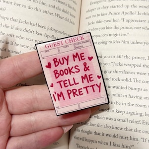 May include: A pink guest check-style sticker with the text "BUY ME BOOKS & TELL ME I'M PRETTY" in red. The sticker has a black border and small heart illustrations. The background is a book.