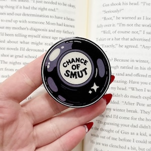 May include: A black and white round pop socket with the text "CHANCE OF SMUT" in a white circle. The pop socket is held in a hand and is set against the backdrop of an open book.