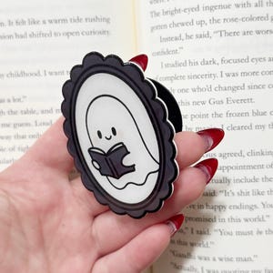May include: A black and white ghost-shaped phone grip with a smiling ghost reading a book. The grip has a scalloped border and is held in a hand, with a book in the background.