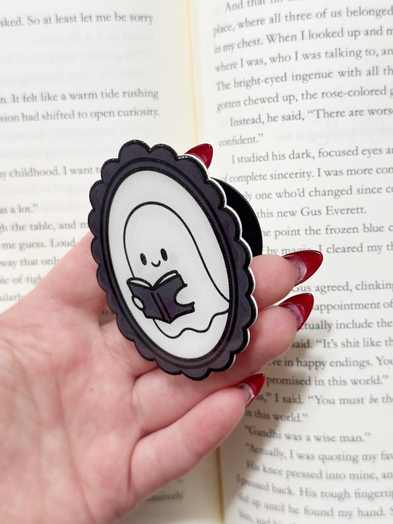 May include: A black and white ghost-shaped phone grip with a smiling ghost reading a book. The grip has a scalloped border and is held in a hand, with a book in the background.