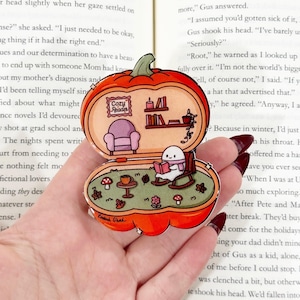 May include: A pumpkin-shaped enamel pin featuring an open interior scene. The top half shows a purple armchair and shelves with books and a candle. The bottom half depicts a white ghost in a rocking chair reading a book, with mushrooms and leaves.