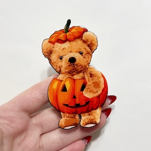 May include: A brown teddy bear wearing an orange pumpkin costume with a carved face. The bear has a pumpkin hat and is standing inside a carved pumpkin. The image is a Halloween-themed design.