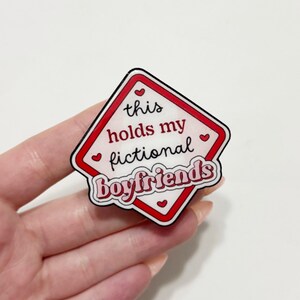May include: A diamond-shaped pin with a red border and the text "this holds my fictional boyfriends" in a playful font. The pin has a white background with red hearts.